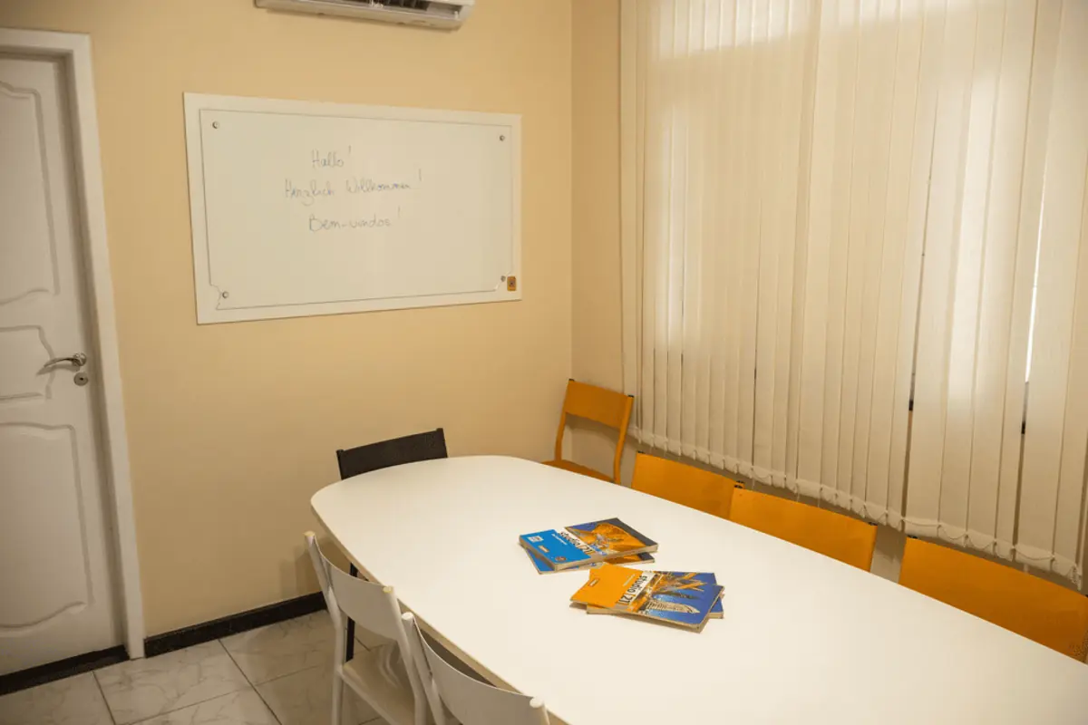 Sala de aula da KONTAKT, com mesa oval, cadeiras laranja e quadro branco escrito “Hallo! Herzlich Willkommen! Bem-vindos!”